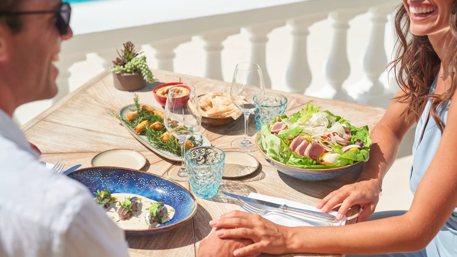 Couple smiling, dining on salad niçoise and other dishes beside white railing