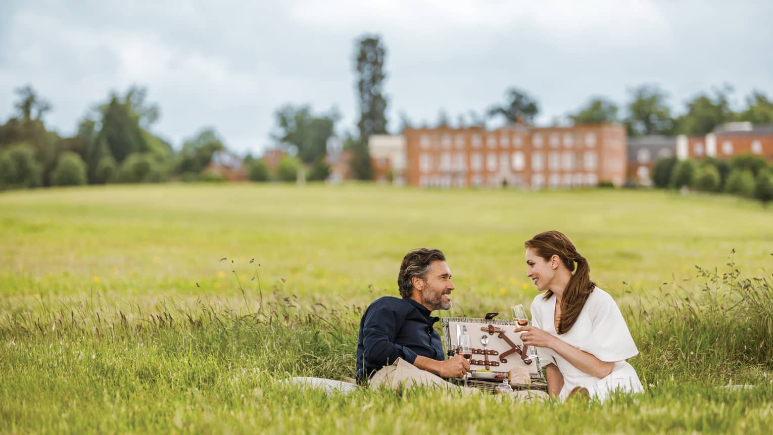 Couple with wine glasses lies in tall green grass with open picnic basket, hotel manor in background