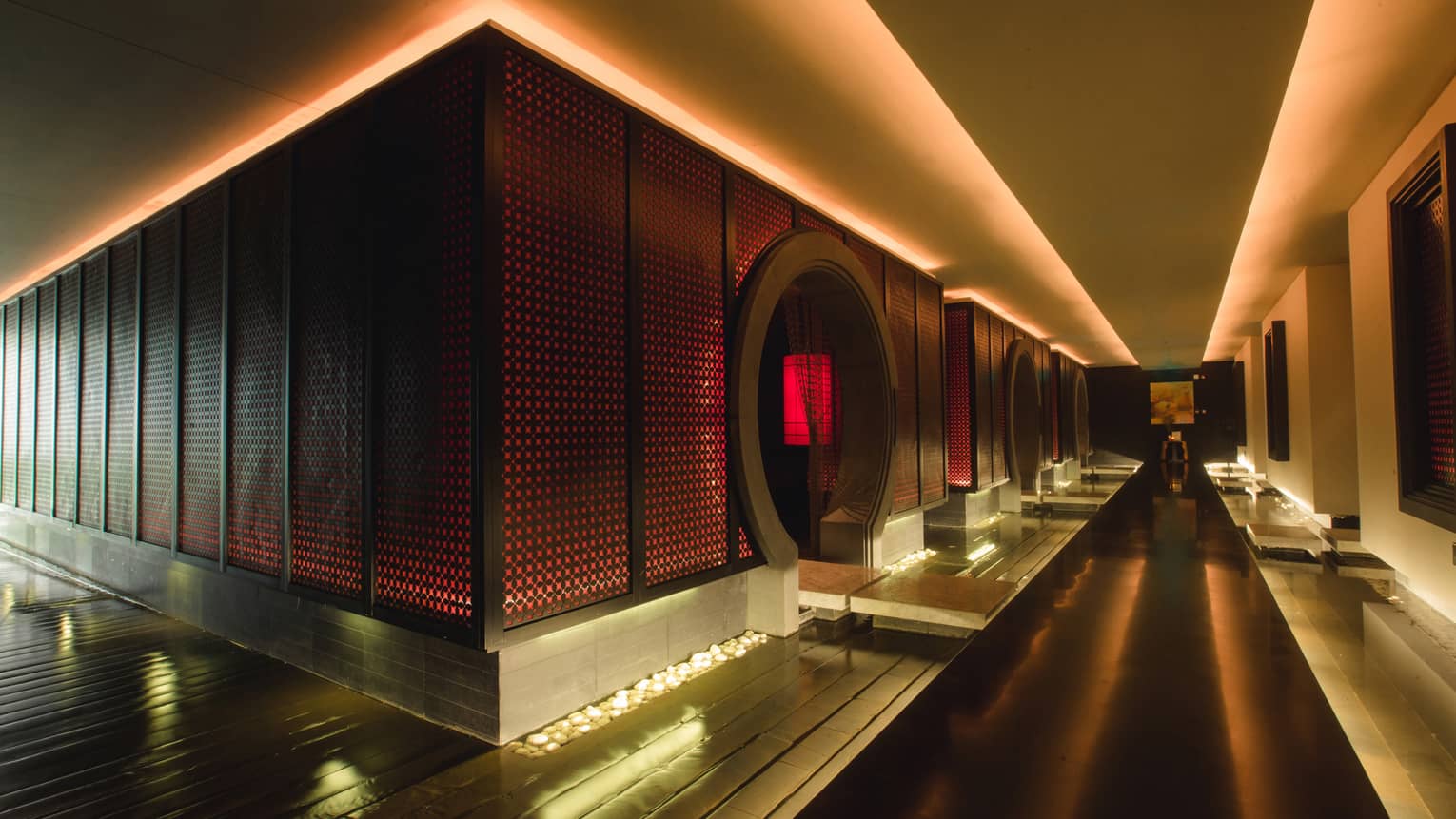 Dark floating walkway down dramatic spa hall past wall with red lights, circular doorways