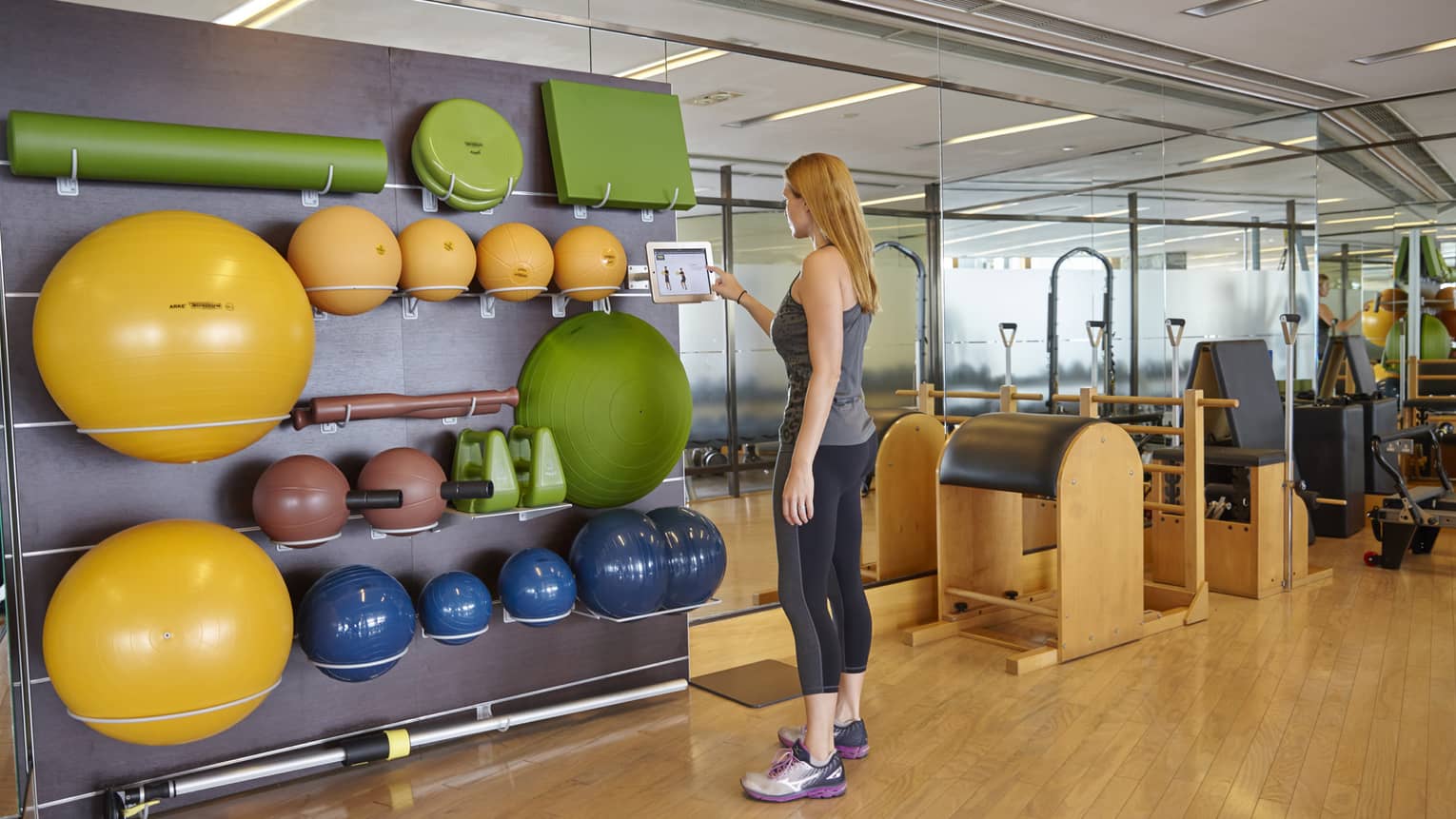 Fitness Centre, woman in workout gear taps taps touch screen by wall with colourful pilates balls, weights 