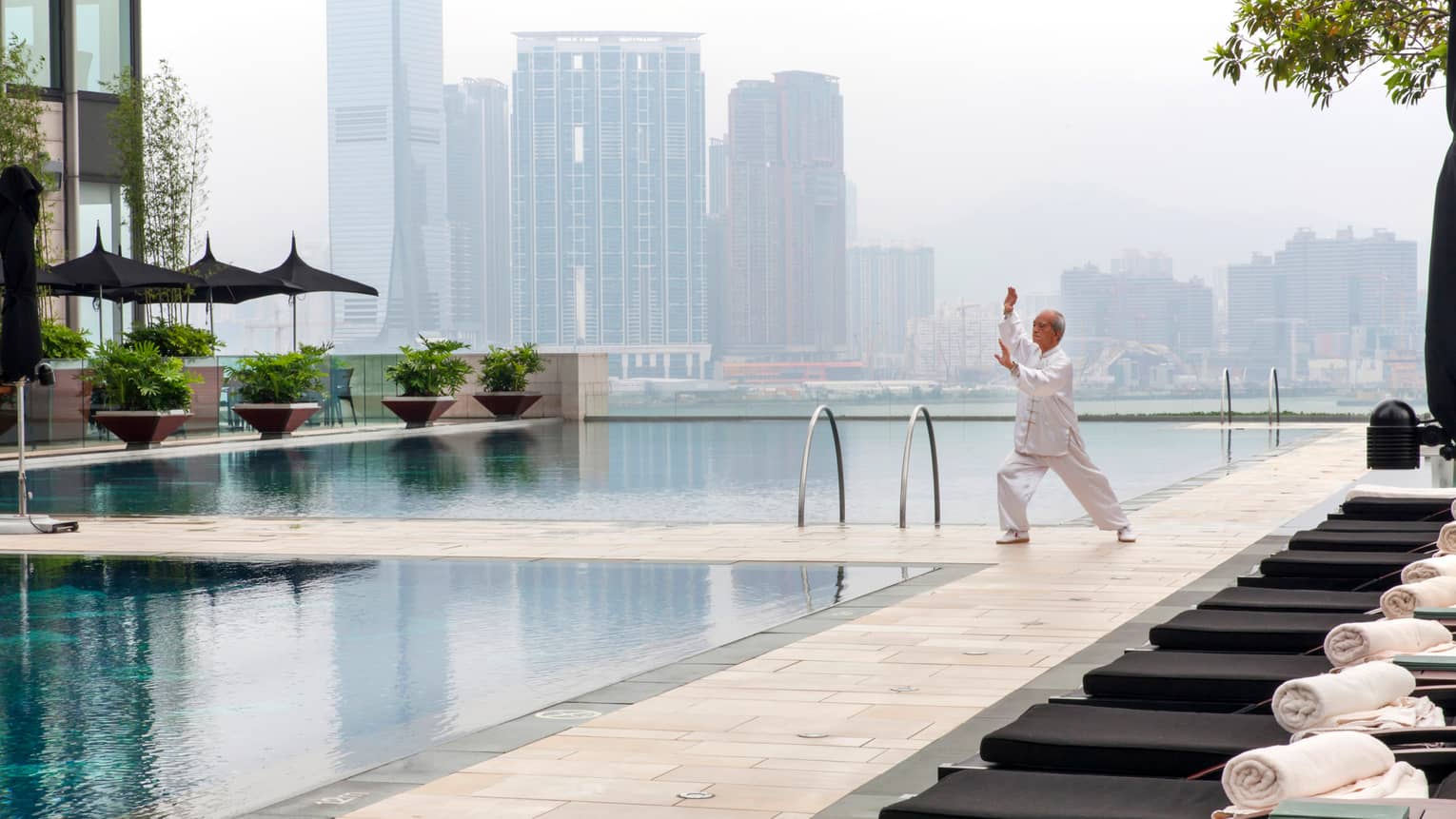 Tai Chi master wearing white holds pose on patio deck by swimming pools, misty skyline