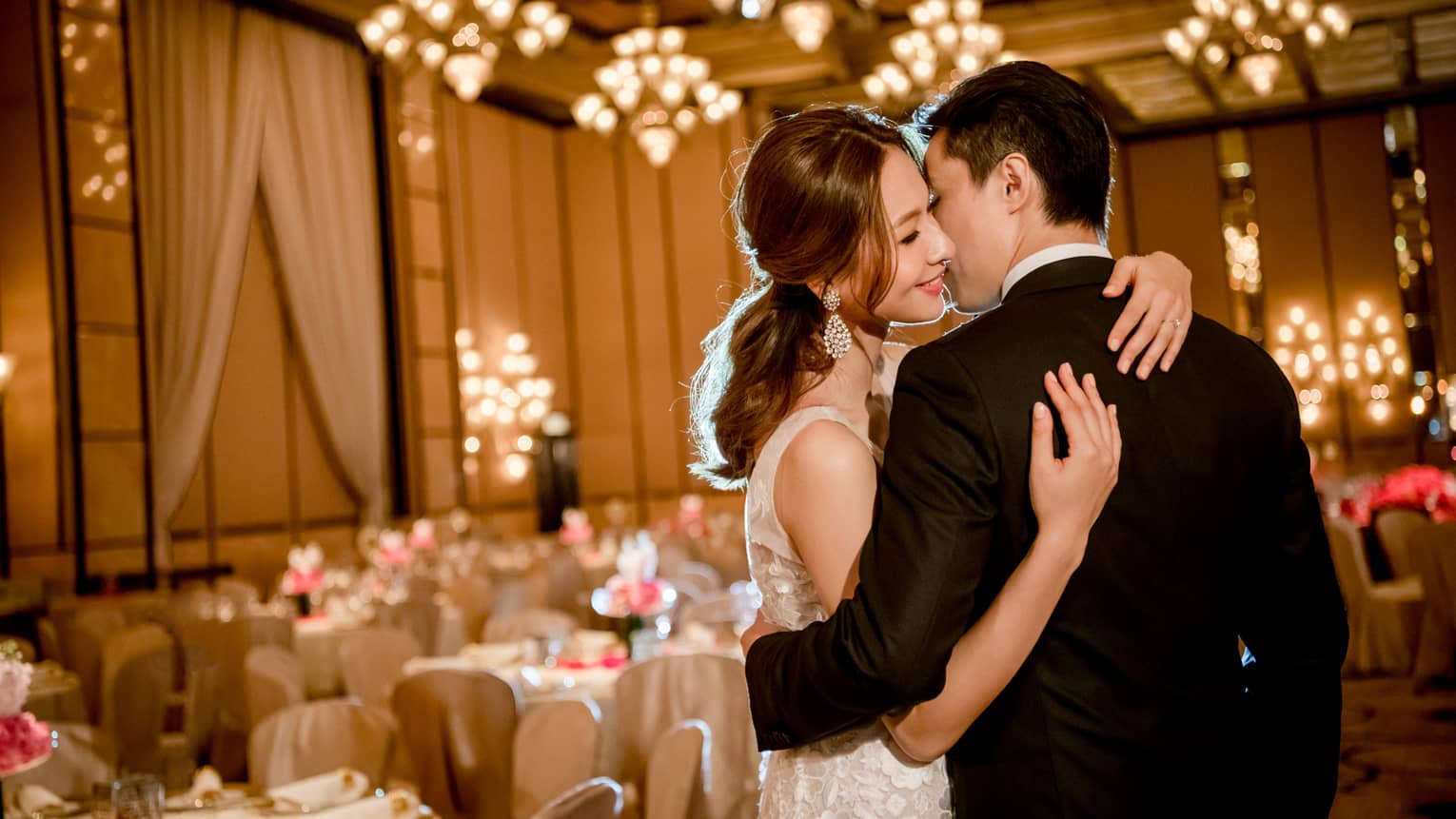 Bride and groom kiss, embrace in elegant ballroom with wedding reception banquet tables