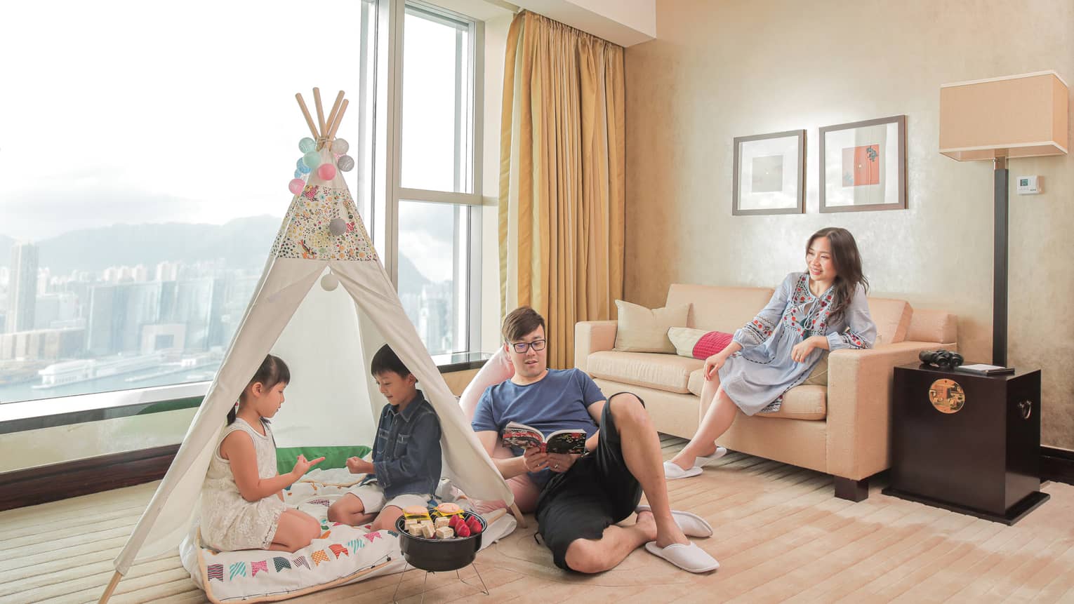 In a brightly lit beige-coloured suite with floor-to-ceiling windows, mom sits on the sofa and looks on as dad reads a book on the floor next to two children playing rock, paper, scissors in a glamping adventure tent set-up with small barbecue in front