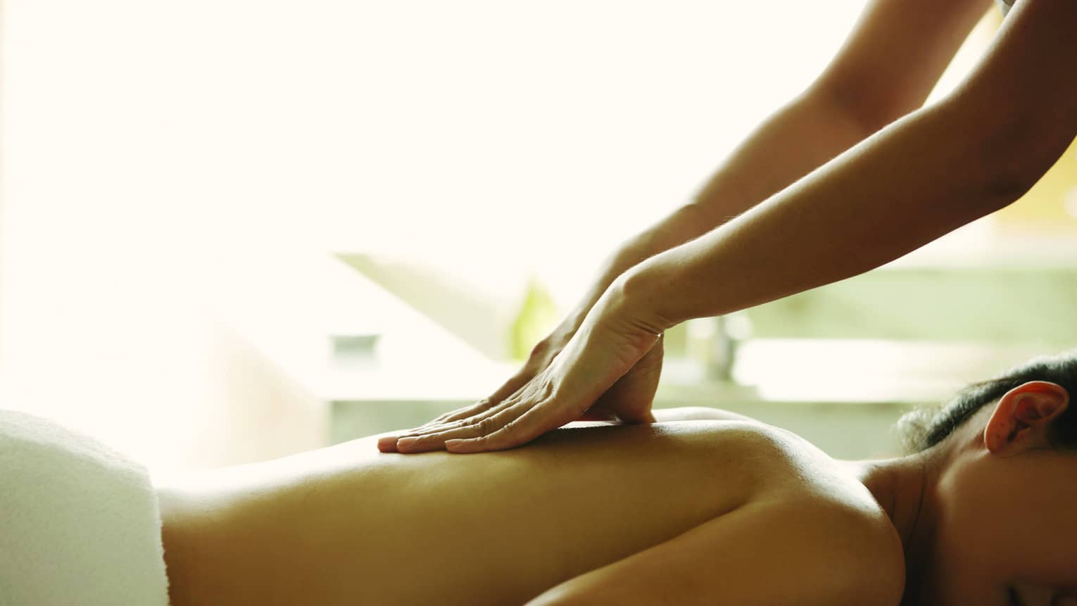 Masseuse reaches over, massages woman's bare back as she lies on spa table