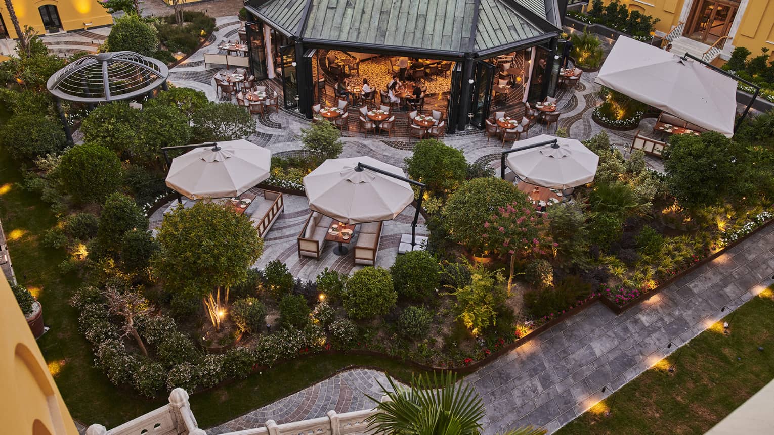 Hotel exterior dining terrace at dusk with covered tables and greenery, lighted walkway