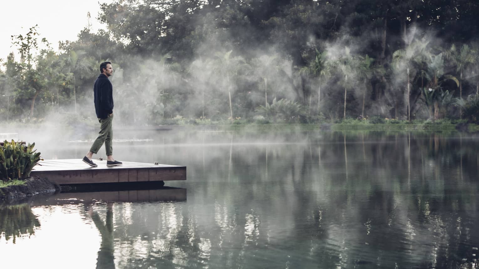 Man walks down a dock as steam rises off the water's surface, his reflection clearly visible 