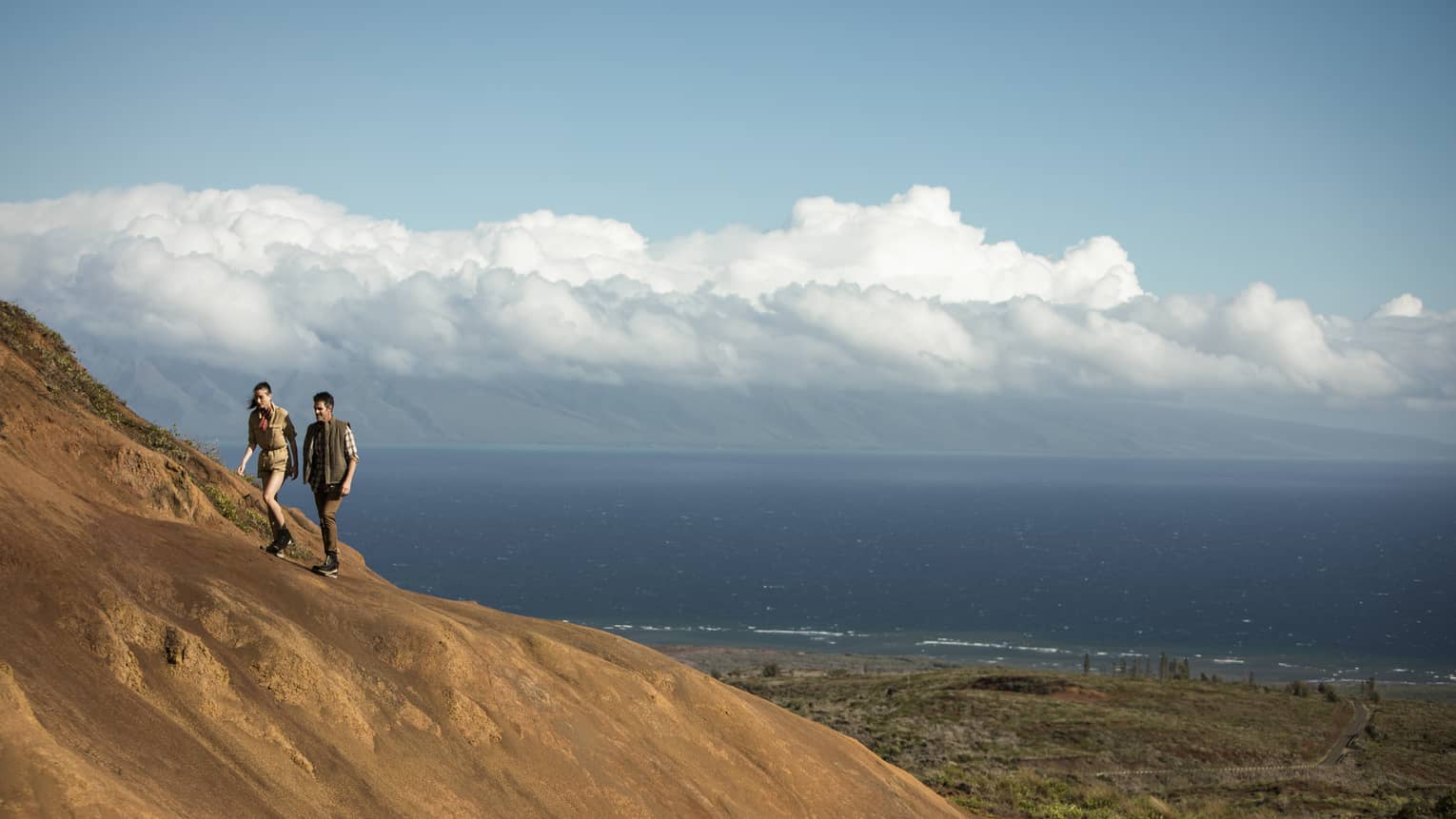 Two hikers climb a large sand dune, puffy white clouds and the ocean looming in the background