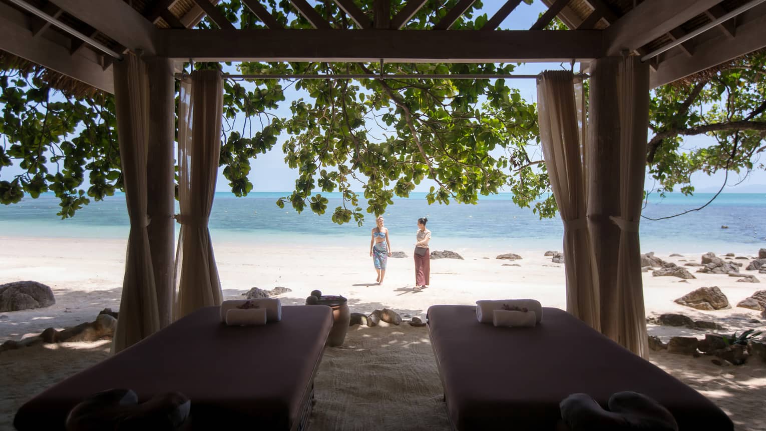 Couples massage beds under cabana in front of two women walking on white sand beach