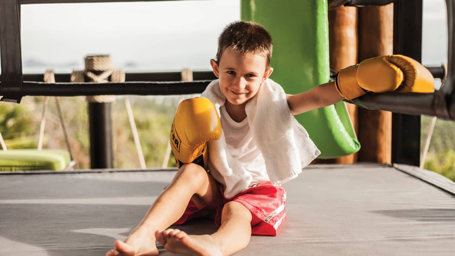Smiling young boy with white towel around neck, boxing gloves sits in sunny outdoor ring