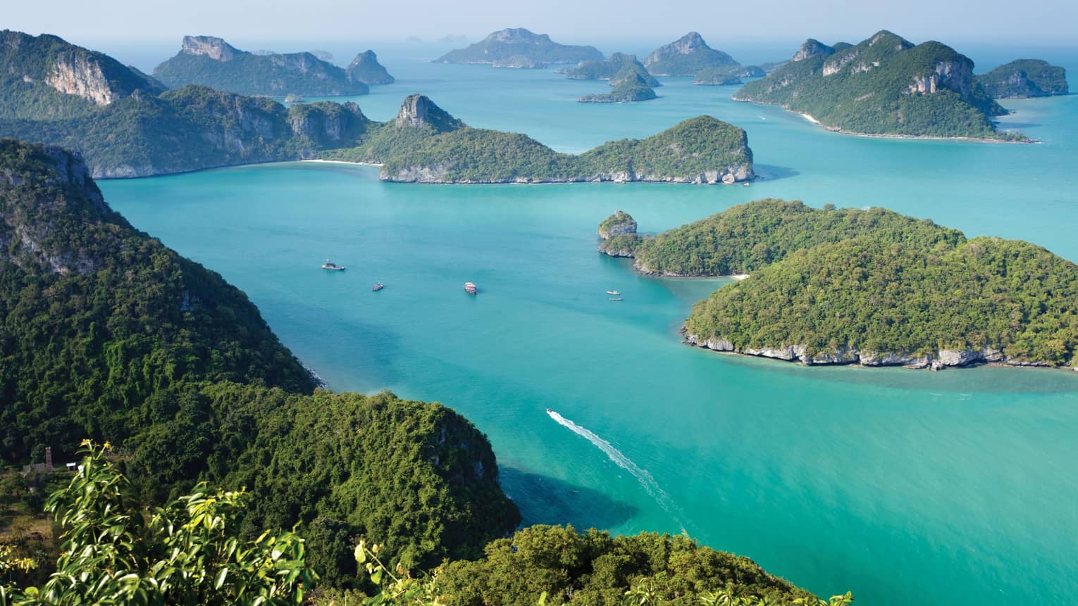 Aerial view of a dozen lush, forested, hilly islands of different sizes surrounded by calm turquoise water dotted with boats.