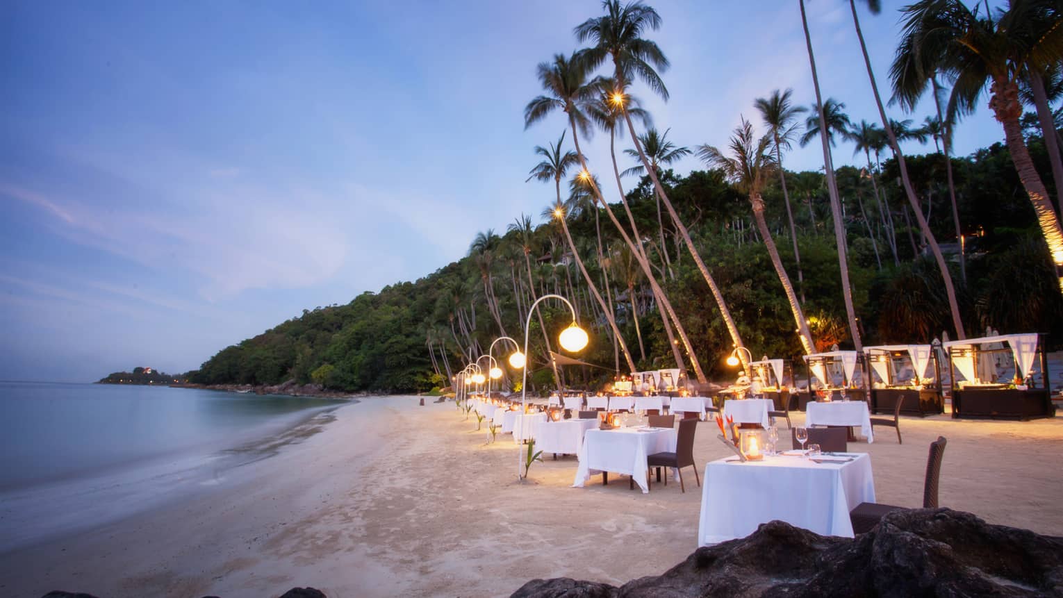 Dining tables set up along white sand beach under palm trees at dusk