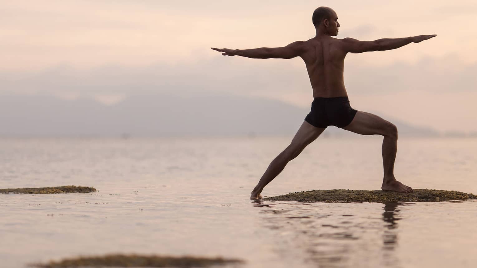 Silhouette of man in swimsuit doing yoga pose on sandbar in ocean at sunset 