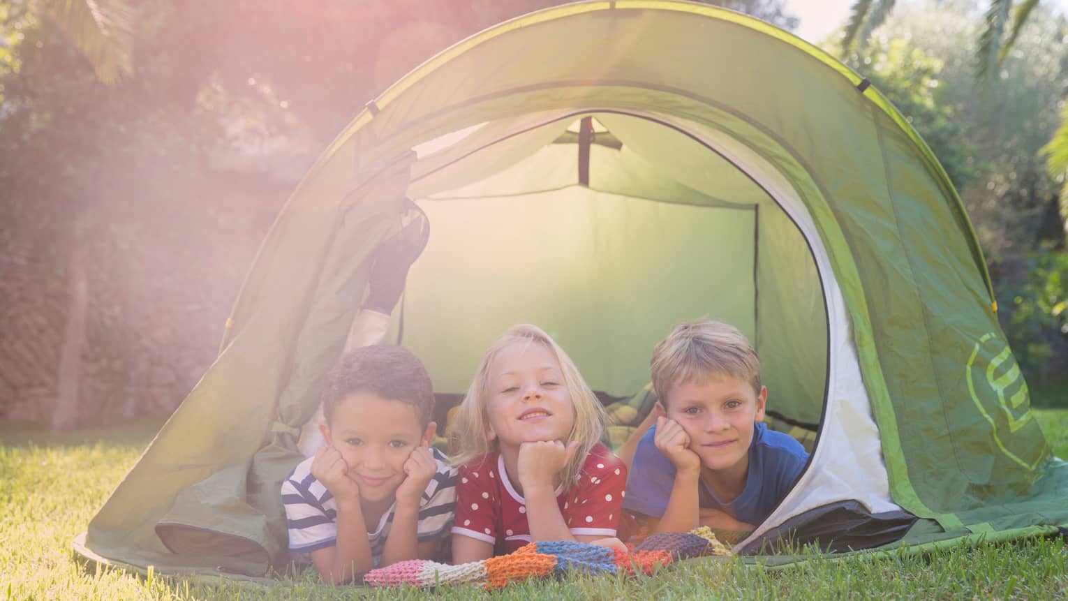 Children lay in a row under small tent opening on grass 