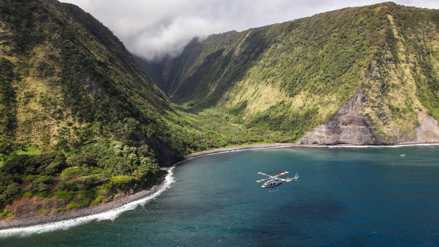 Helicopter flies over ocean by sweeping green volcanic mountains