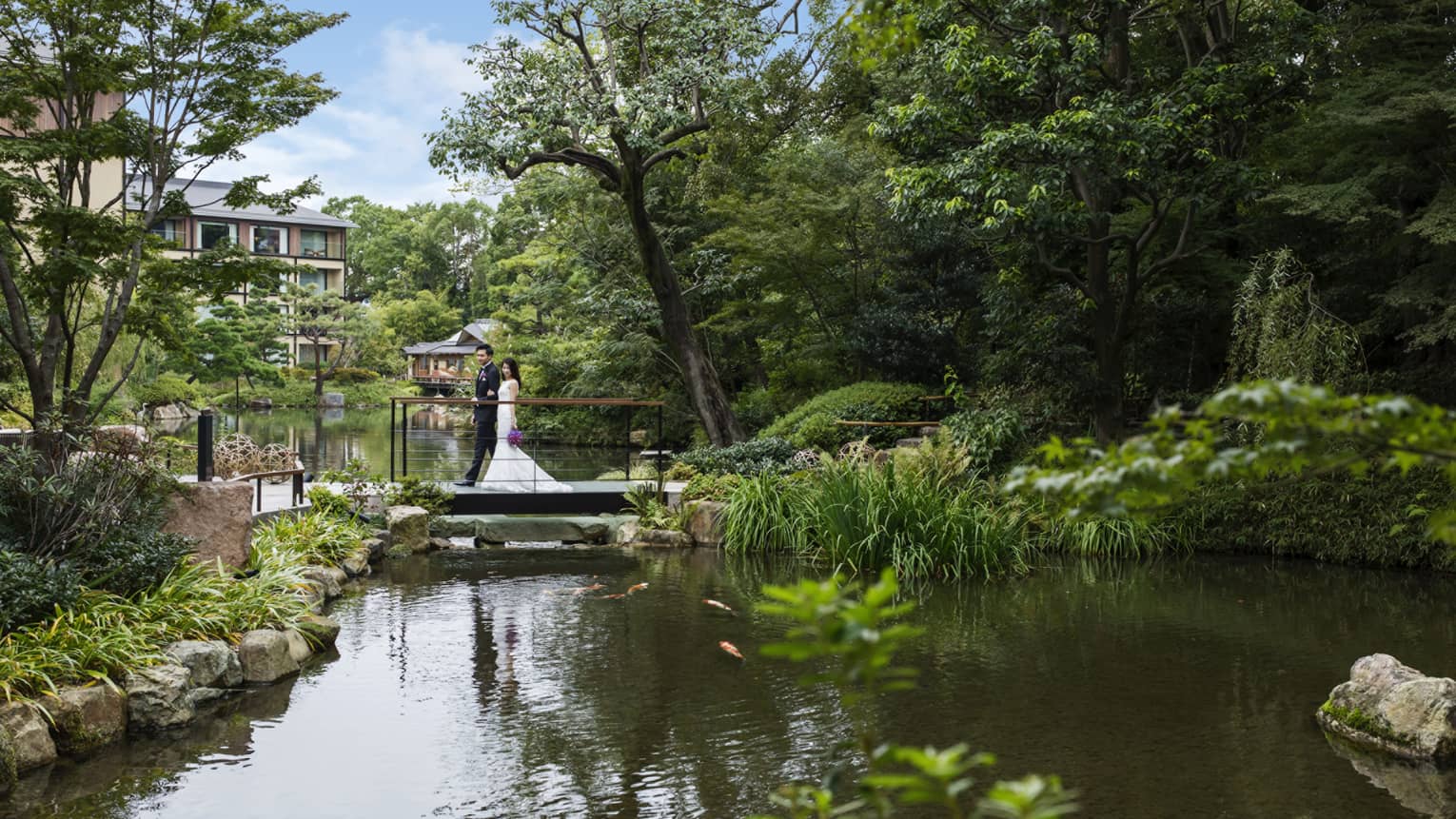 Bride and groom walk across small bridge over pond under trees
