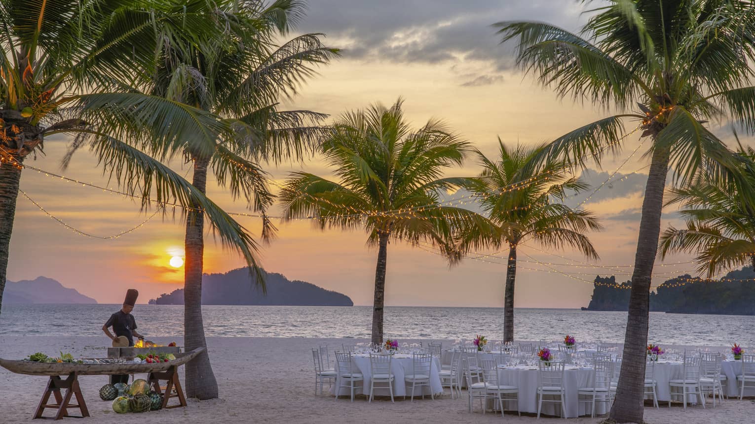 Tables set for dinner on the beach under a canopy of palm trees, a chef cooking over an open flame against the setting sun.