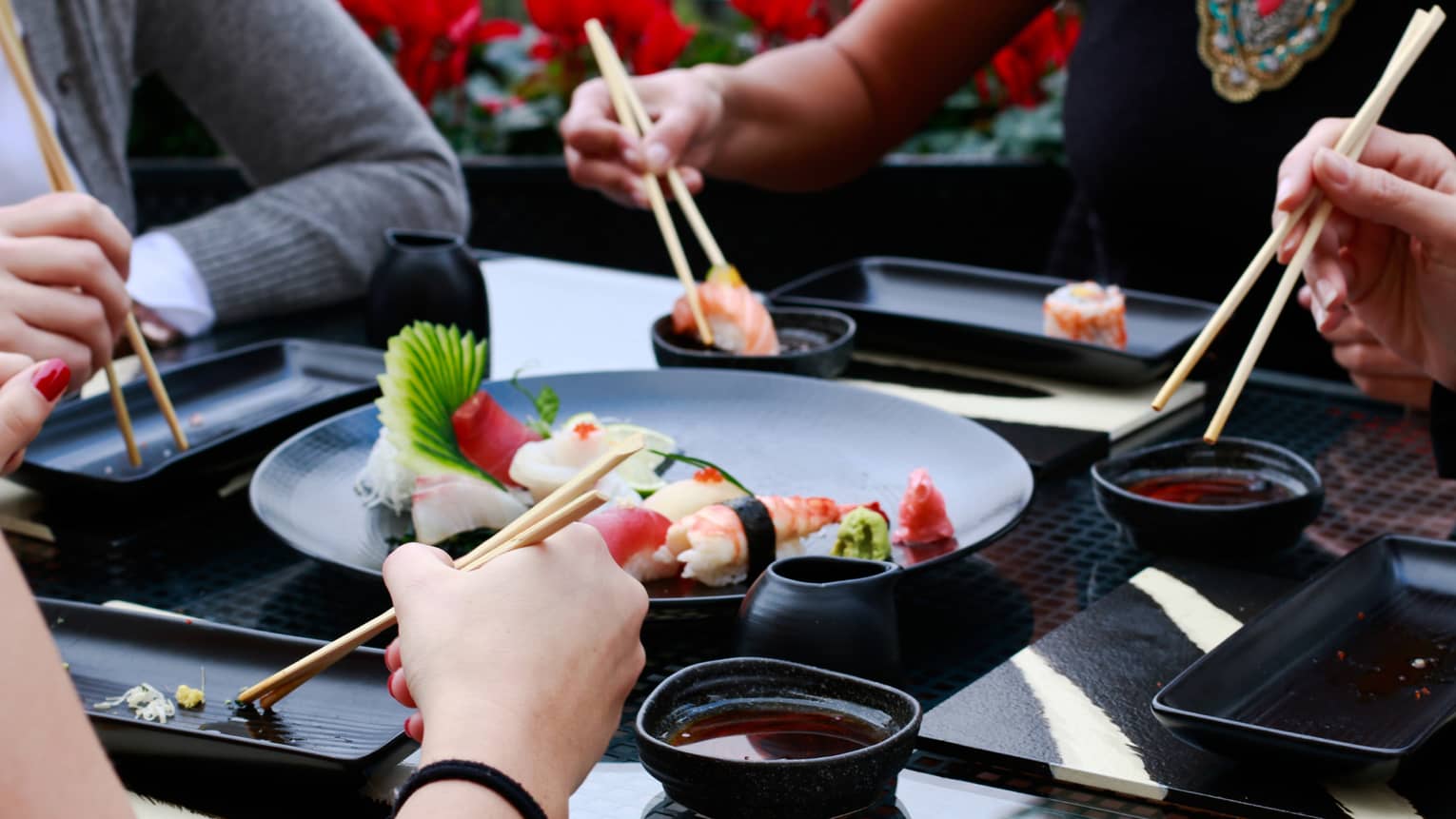 Close-up of four sets of hands holding chopsticks around black plate with sushi rolls, bowls of soy sauce