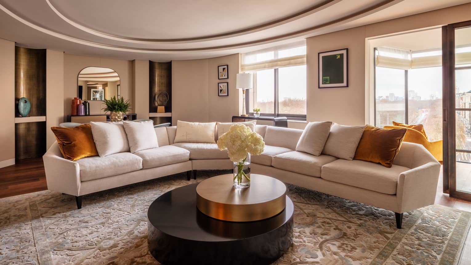 The Hyde Park Suite at Four Seasons London Park Lane in Mayfair showcases a sophisticated living room with a panoramic view of the city. The room features a long, sleek white sofas with orange throw pillows, arranged around a large circular black coffee table with a gold rim. The area is adorned with a luxurious beige patterned rug, adding texture and warmth. Artworks hang on the walls, and a large window frames the stunning view outside, providing ample natural light. The decor is elegantly completed with a neutral colour palette and modern furnishings, creating an inviting and stylish space.