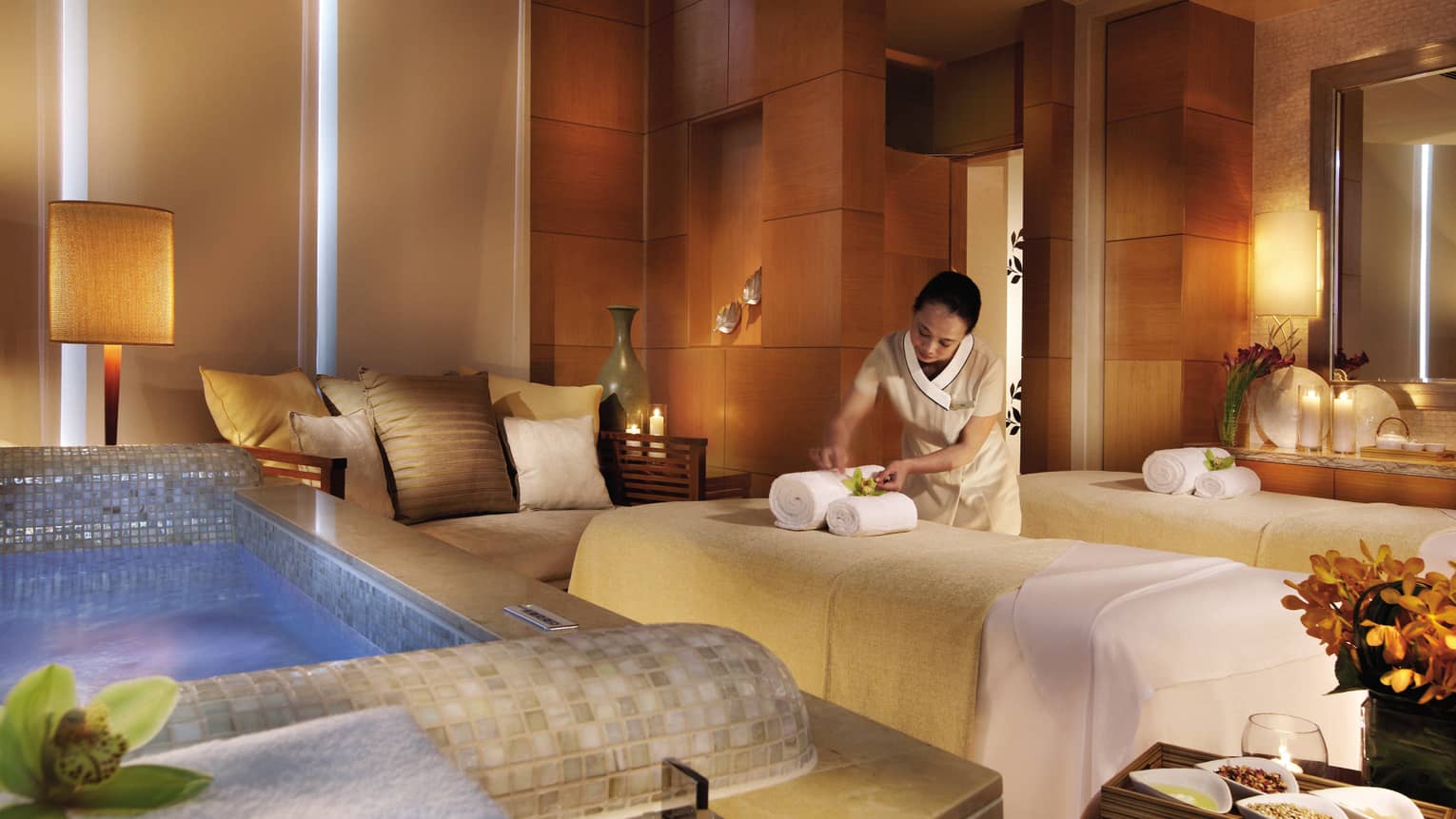 Spa staff sets rolled towels on massage bed near small sofa, tile bathtub in Couples Suite