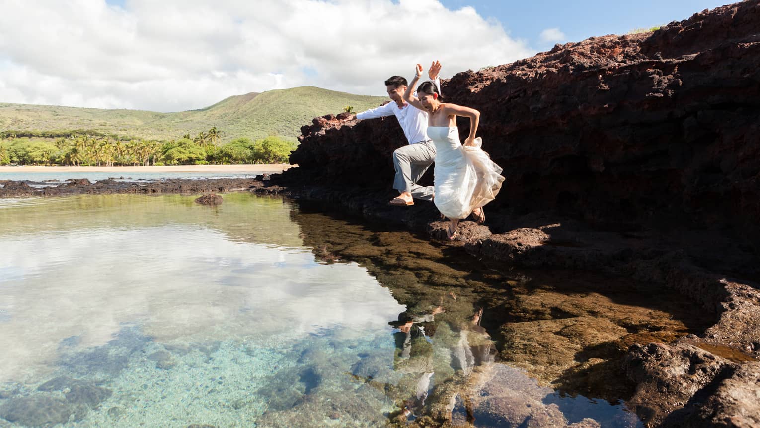 Bride and groom jump into clear lagoon under lava rock cliff