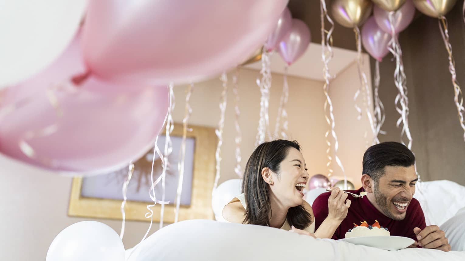 Laughing couple share a dessert in bed surrounded by pink, gold and white balloons