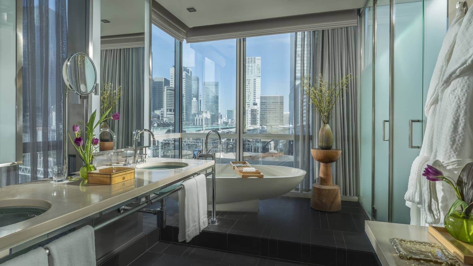 Bathroom with raised standalone tub next to a floor-to-ceiling window, double vanity and frosted glass shower