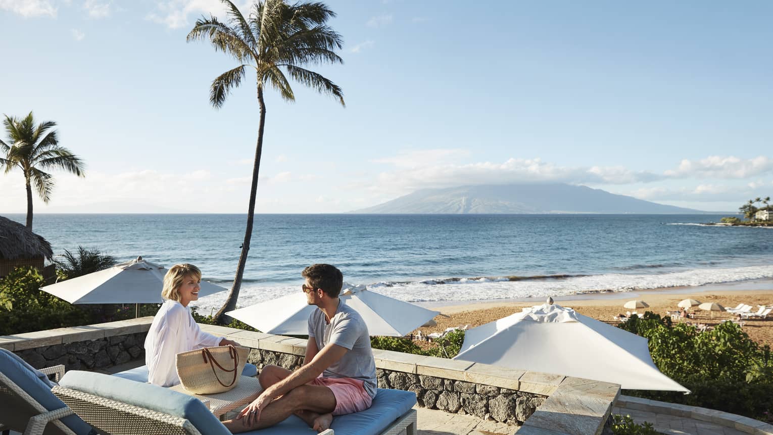 Man in shorts and smiling woman in beach shirt sit on edge of lounge chair, beach and ocean in background