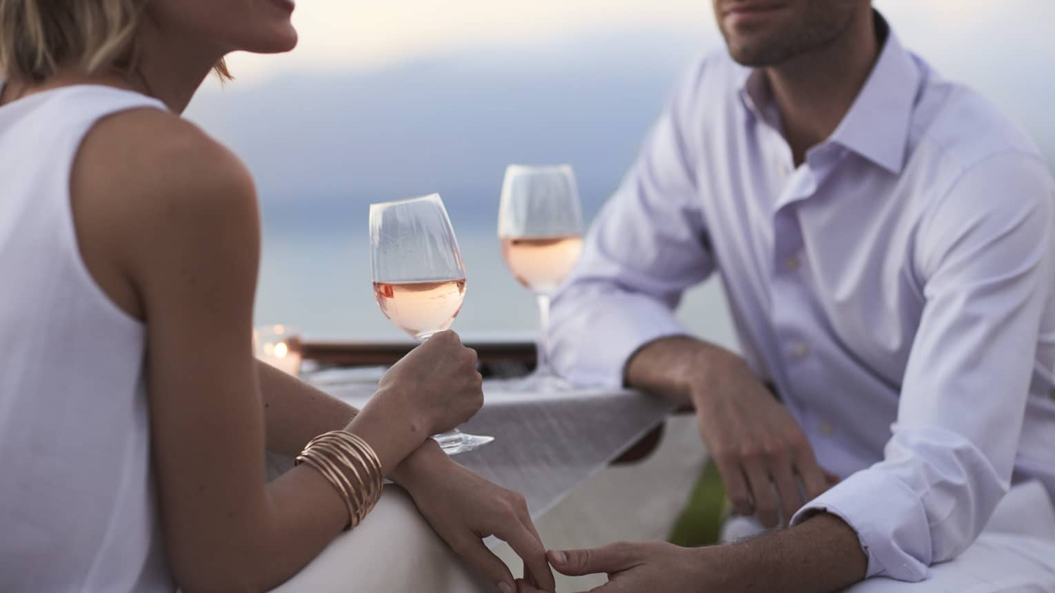 Close-up of woman and man in white, sitting and holding glasses of rose wine at dusk