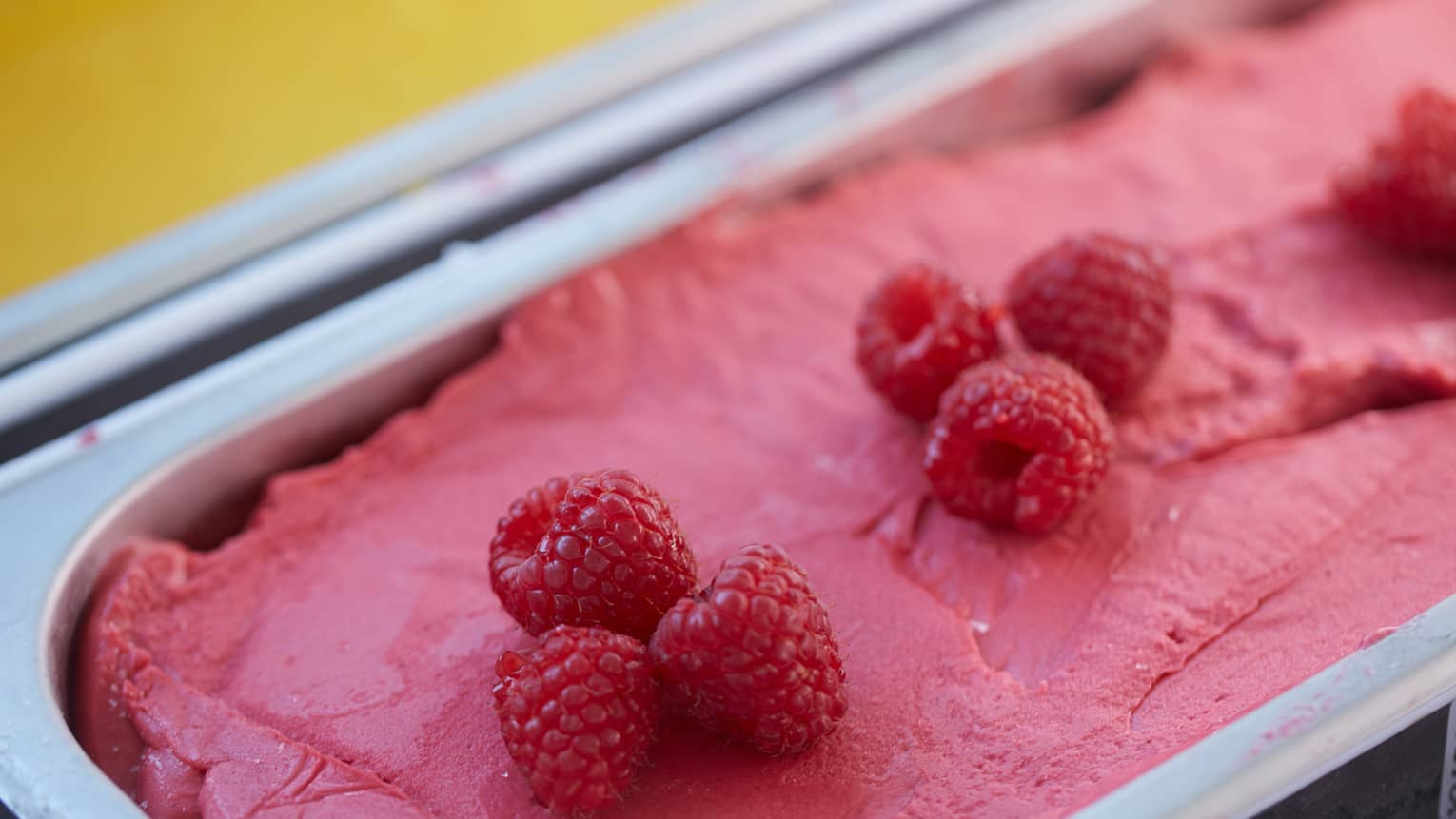 A close-up of raspberry gelato in a metal container, ready for serving, with fresh raspberries on top