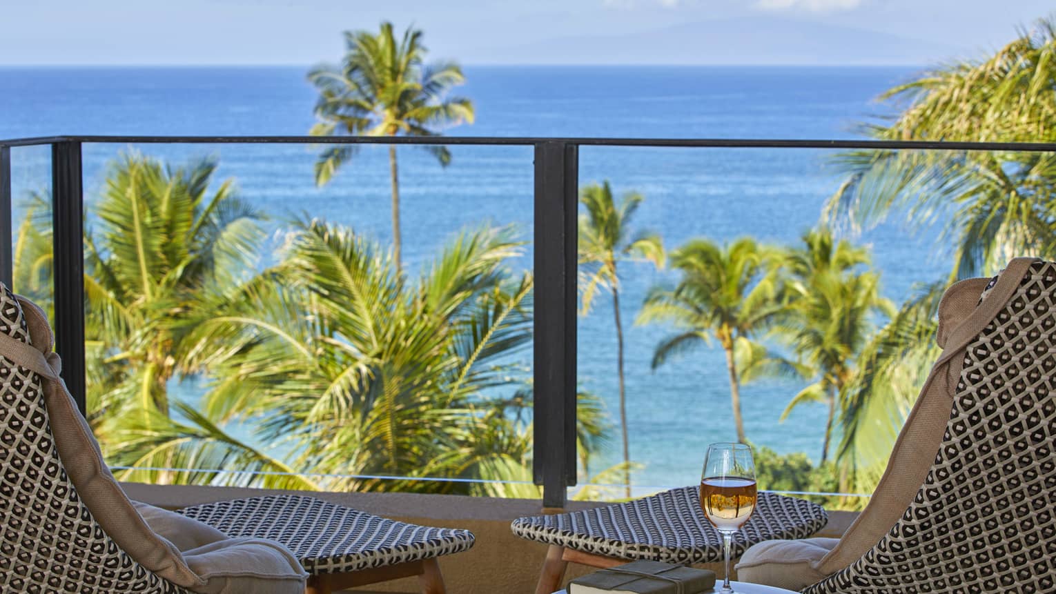 Balcony with rattan chairs, round table with book and glass of wine, overlooking palm trees and blue ocean