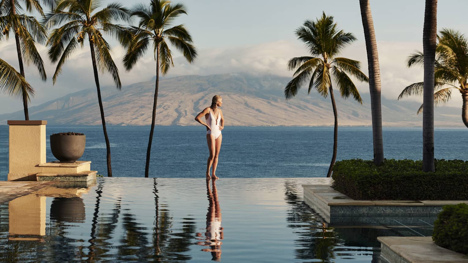 Woman stands at edge of infinity pool, nearby Hawaiian island in distance