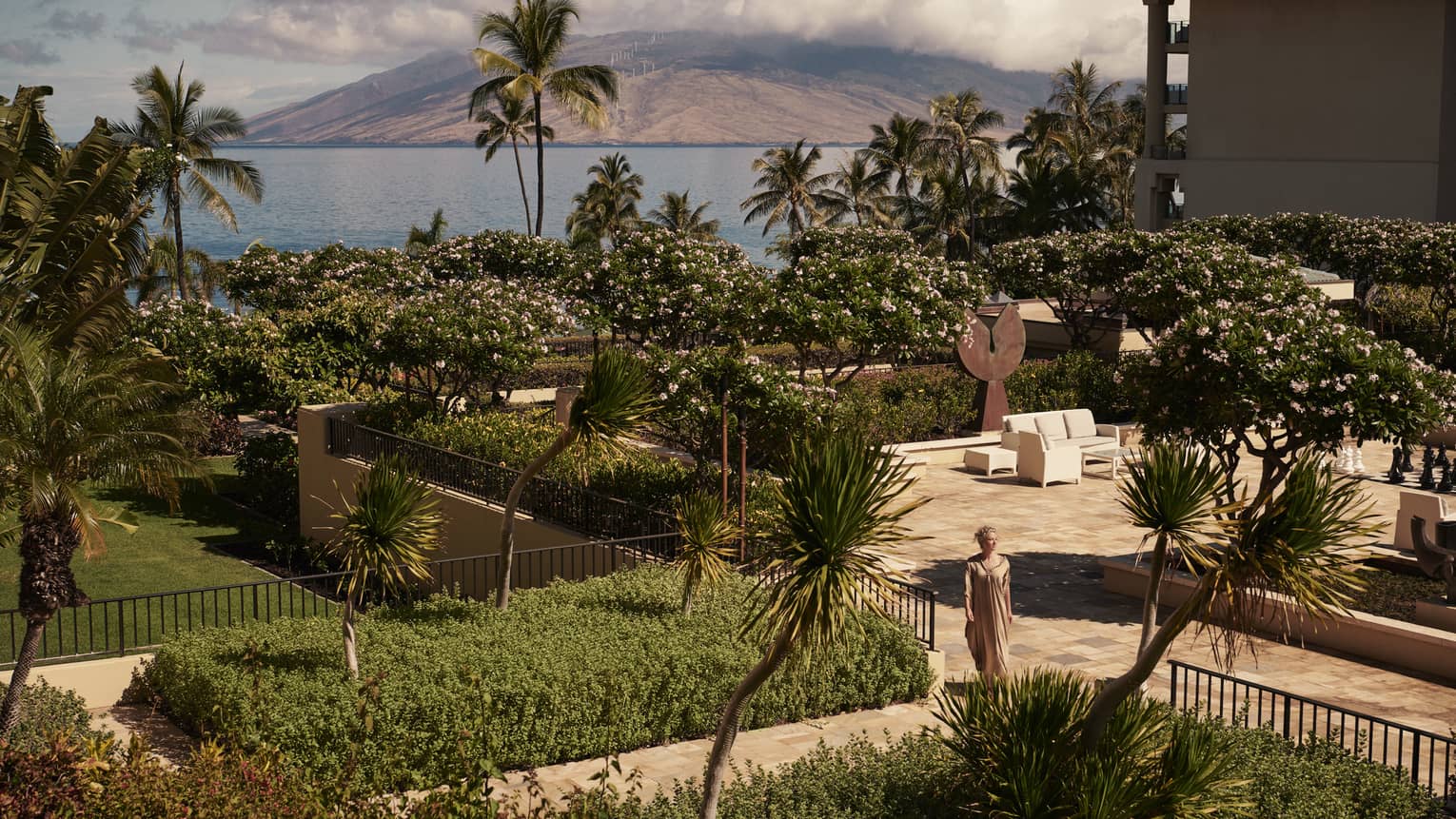 Woman walks through gardens on resort grounds in Maui, ocean in background
