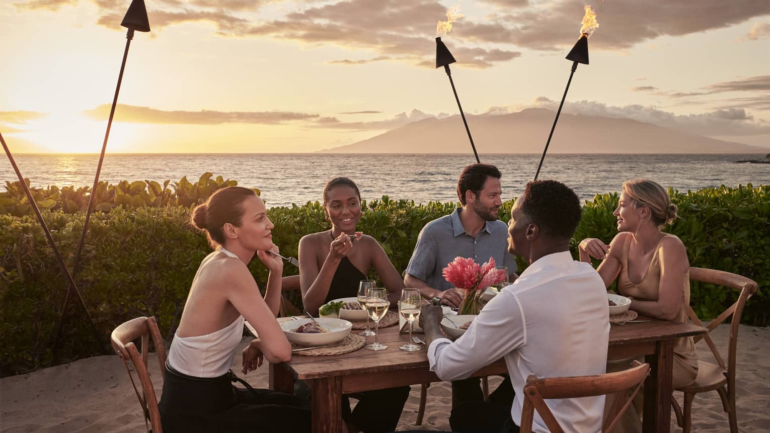 Three women and two men at table on beach, surrounded by tiki torches