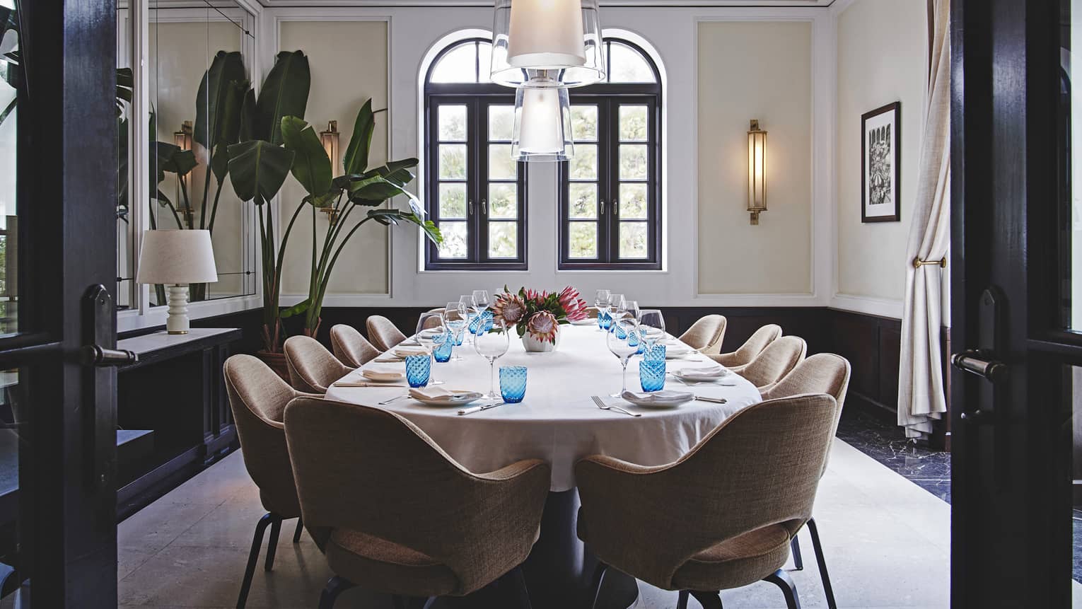A private dining room with a long table surrounded by leather chairs, two lamps hang from the ceiling while tall plants are in the corner next to windows.