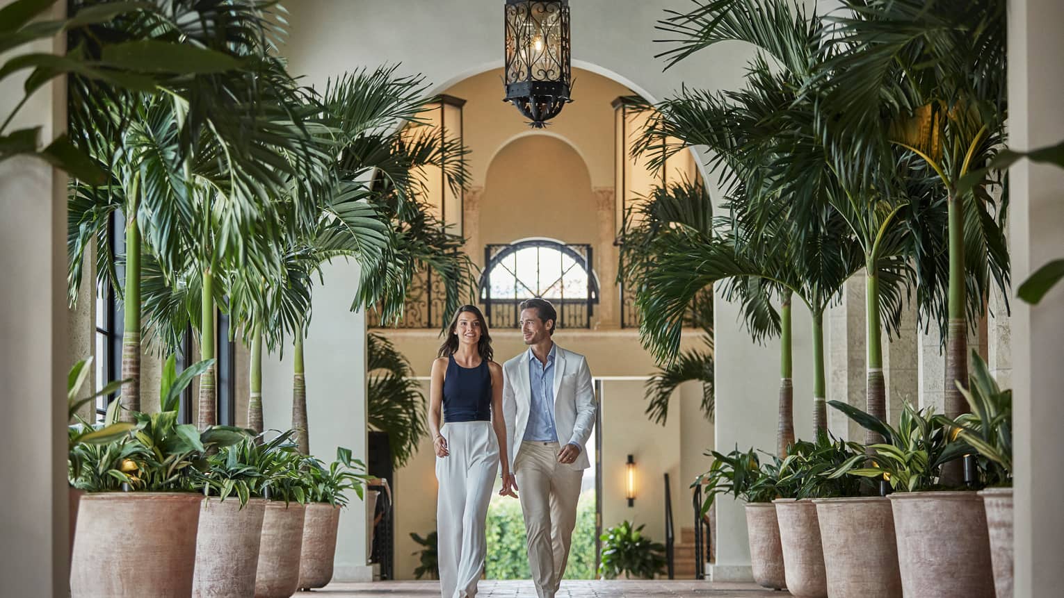 Couple walks down hallway lined with potted palm trees under arched ceiling