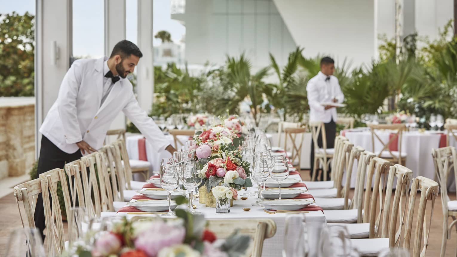 Hotel staff wearing white suit jackets set long wedding dining table with flowers