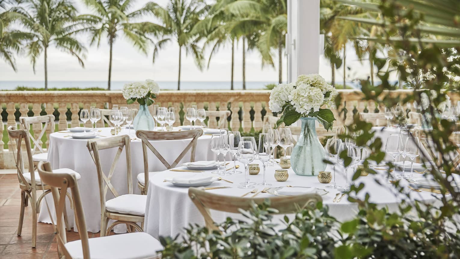 Patio wedding reception dining tables set with glass vases with white flowers, palm trees in background