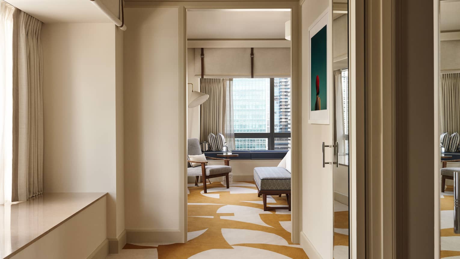View through an open doorway into a luxurious one-bedroom suite at the Four Seasons Hotel in Miami, showcasing a modern living area with stylish furniture, including a plush grey sofa and matching armchairs. The suite features a geometric patterned rug and large windows that provide a sweeping view of the city skyline. The interior design combines contemporary elegance with comfort, highlighted by soft, natural lighting and neutral colour tones.
