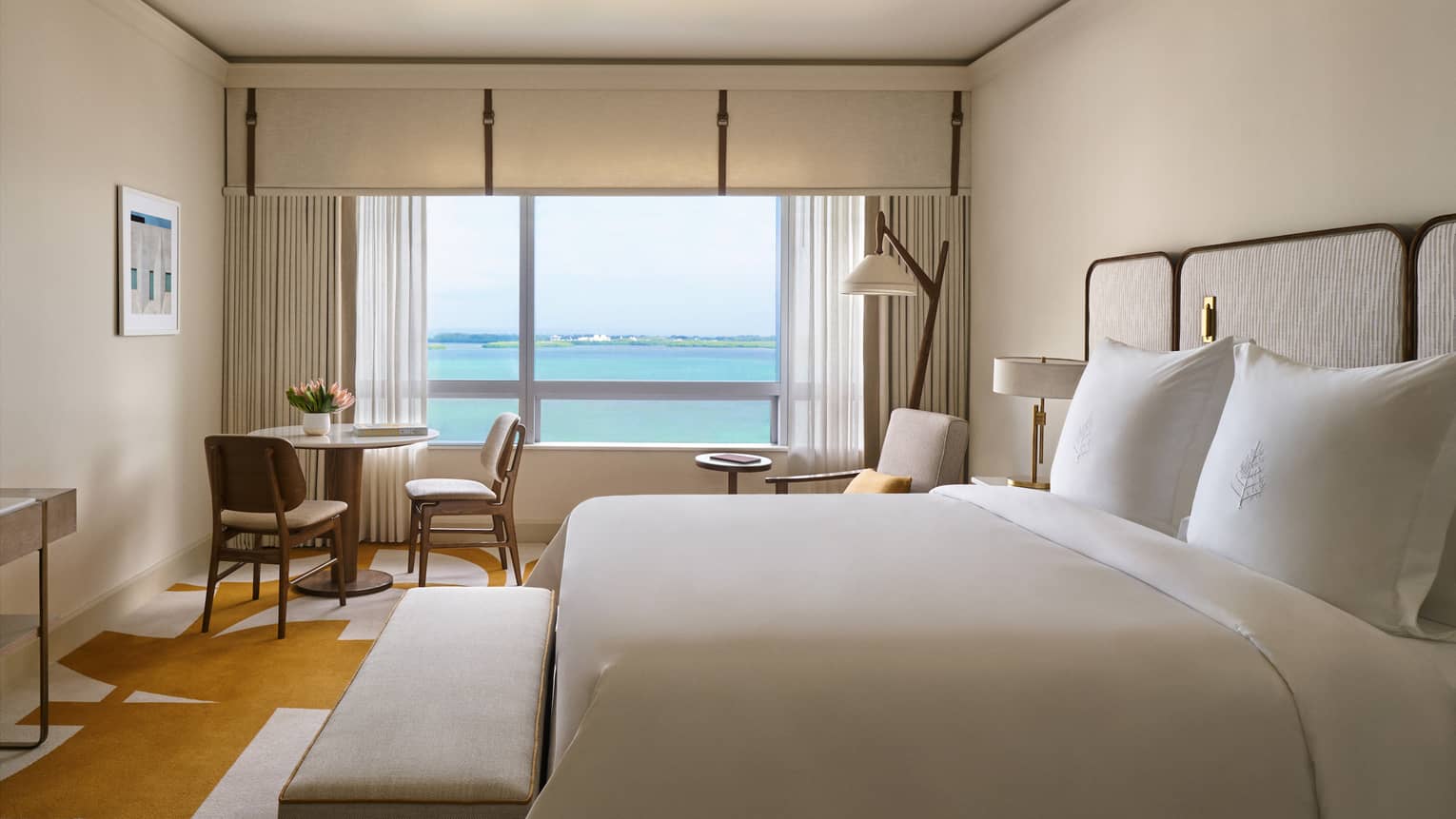 Elegant and serene guest room at the Four Seasons Hotel in Miami, featuring a large bed with white linens and a beige headboard. The room includes a small seating area with two chairs and a table, all bathed in natural light from floor-to-ceiling windows offering a stunning view of the bay.