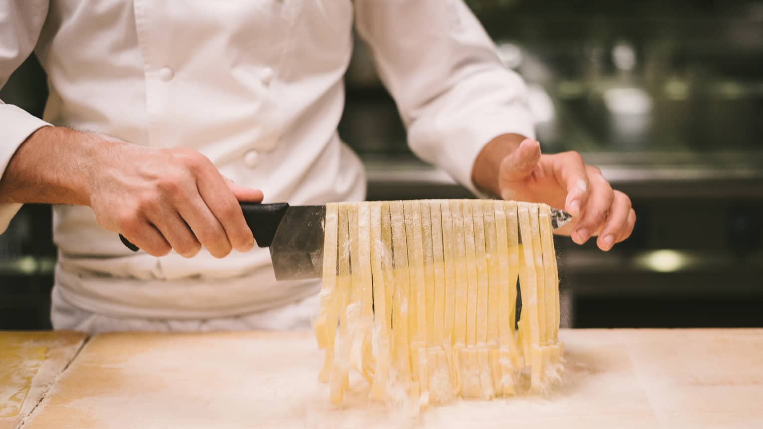 Chest down of chef in white coat holding up chef's knife draped with row of freshly cut pasta ribbons