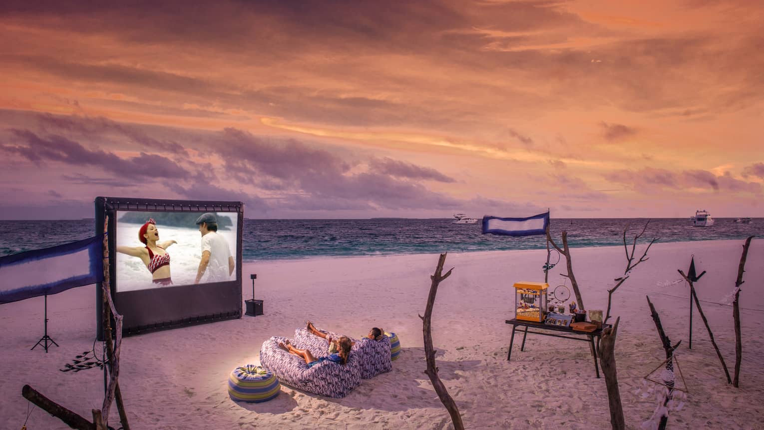 ,Two people in beanbag chairs enjoy a movie night on the beach at dusk, popcorn stand on sand behind them