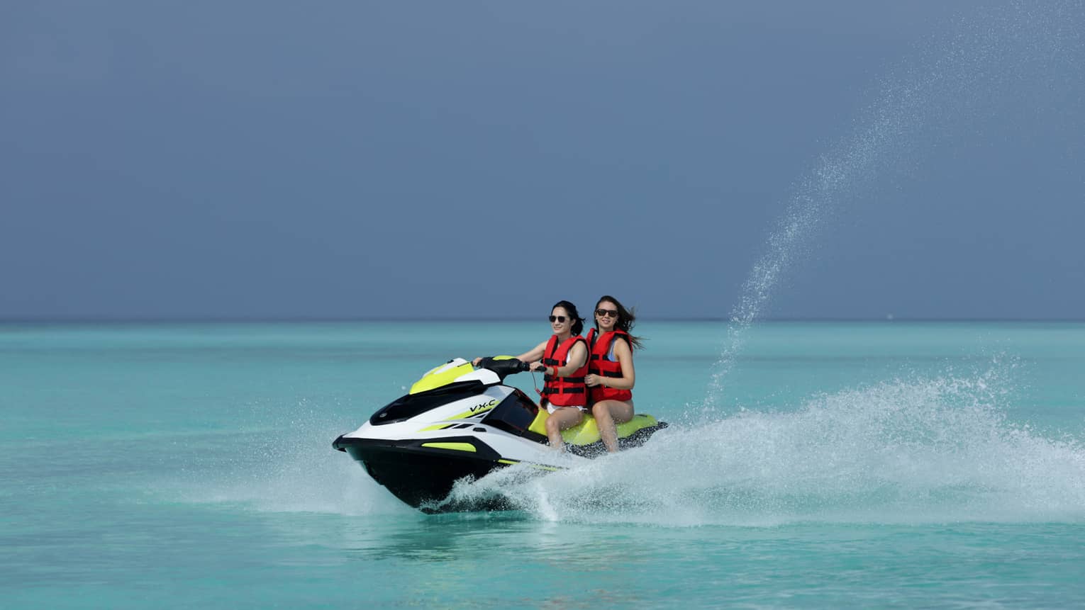 Wearing life jackets, two smiling jet-skiers ride through the clear ocean, water spurting up from beside and behind the boat.