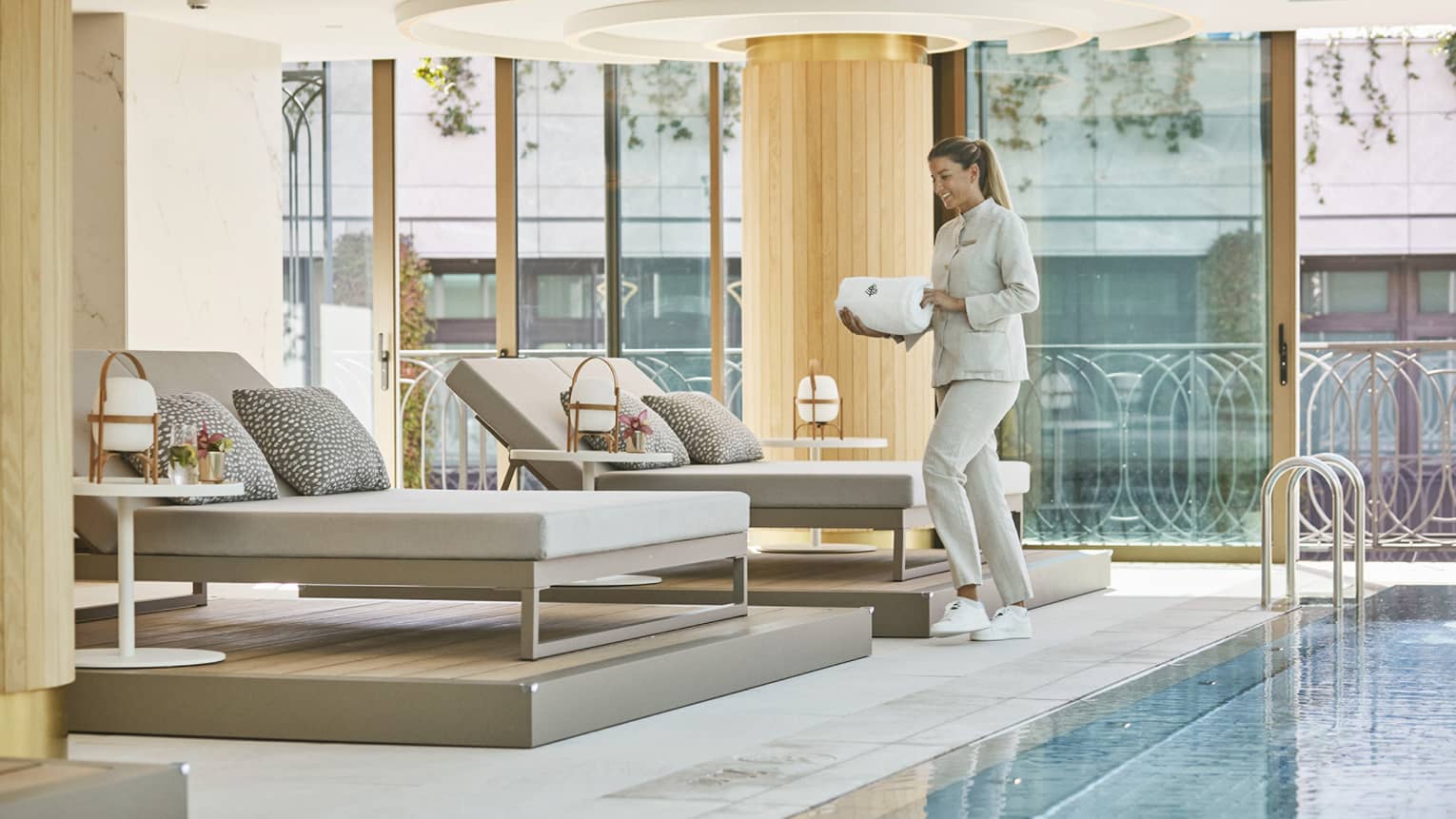 A woman carrying white towels to cushioned lounge seat next to indoor pool