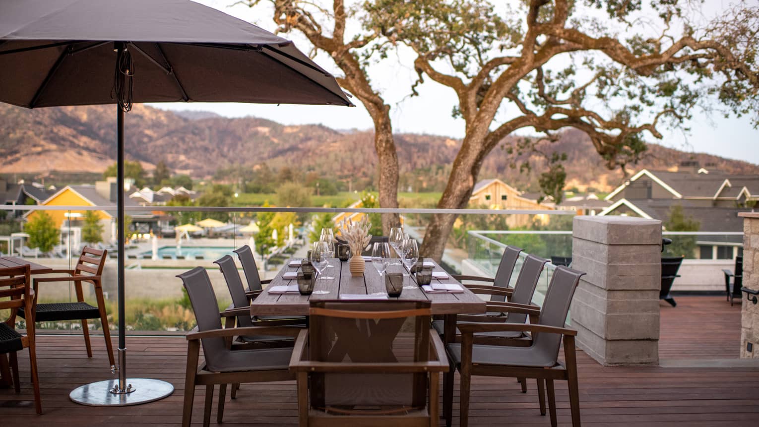 An outdoor eating area near hills and trees with a long rectangular table and umbrellas.