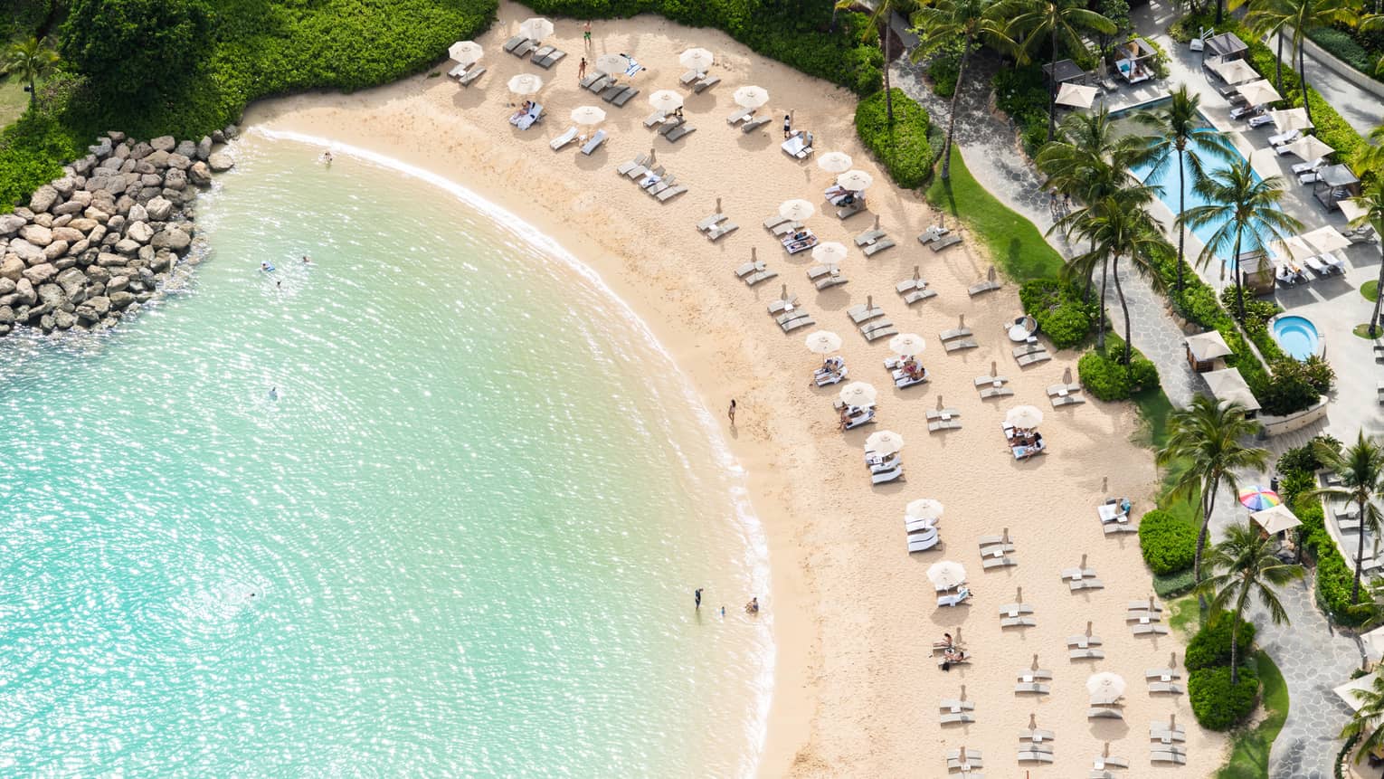 Aerial view of curved beach and clear blue water, beach chairs, resort pool
