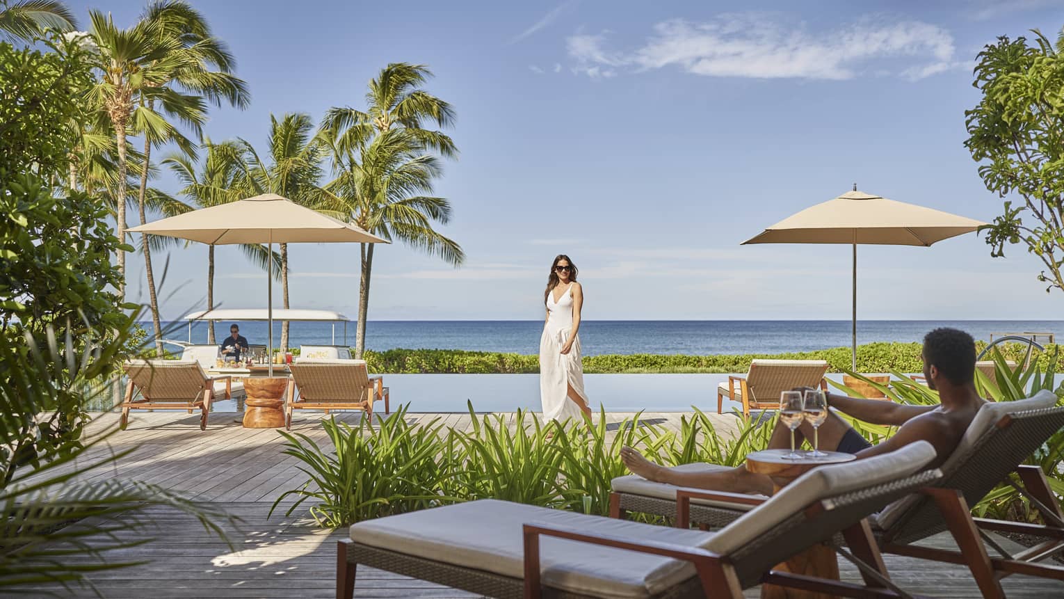 A woman dressed in white walks toward a lounge chair by an outdoor infinity pool