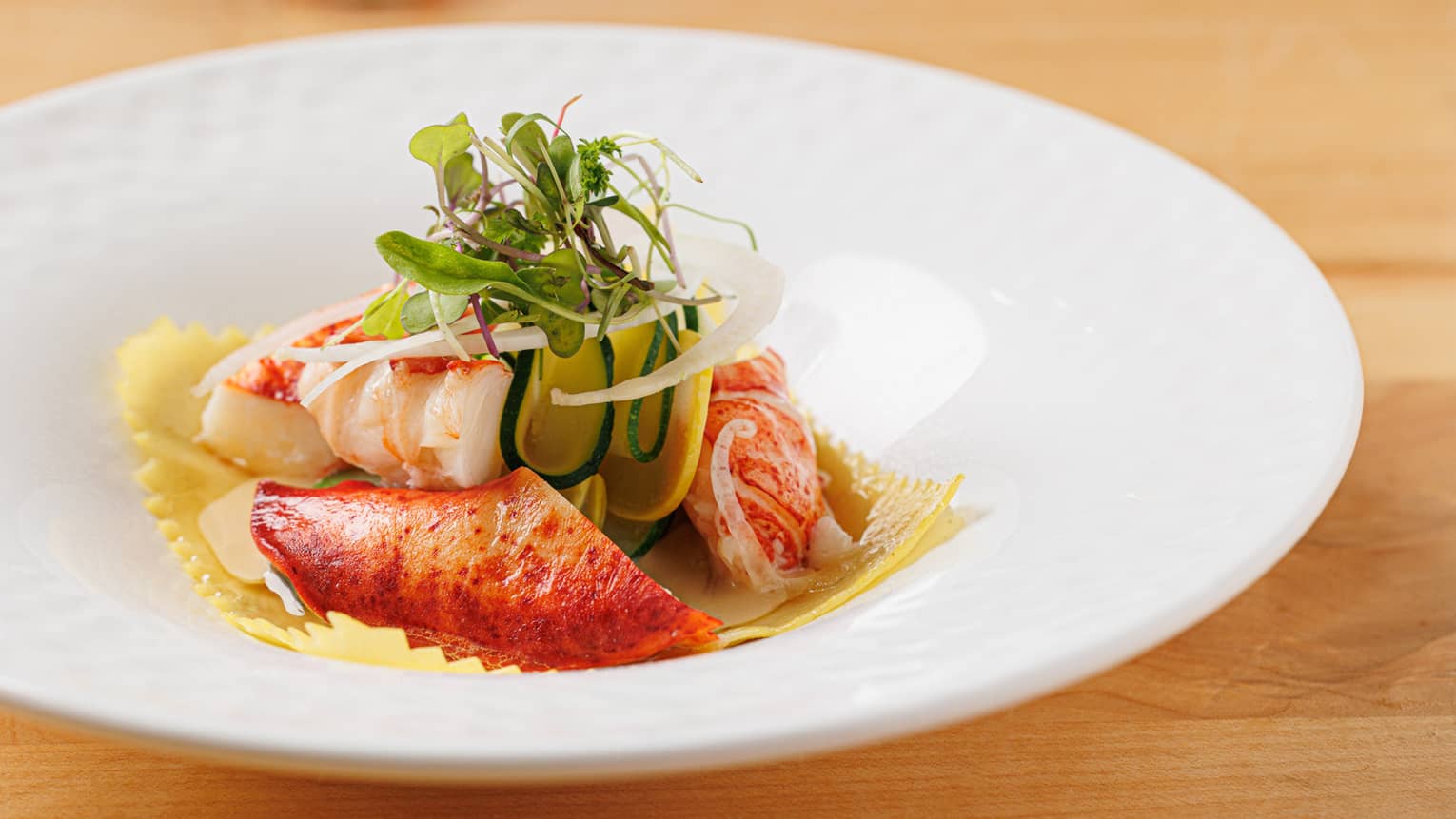 Lobster atop fresh pasta, garnished with micro greens and shaved vegetables in white dish