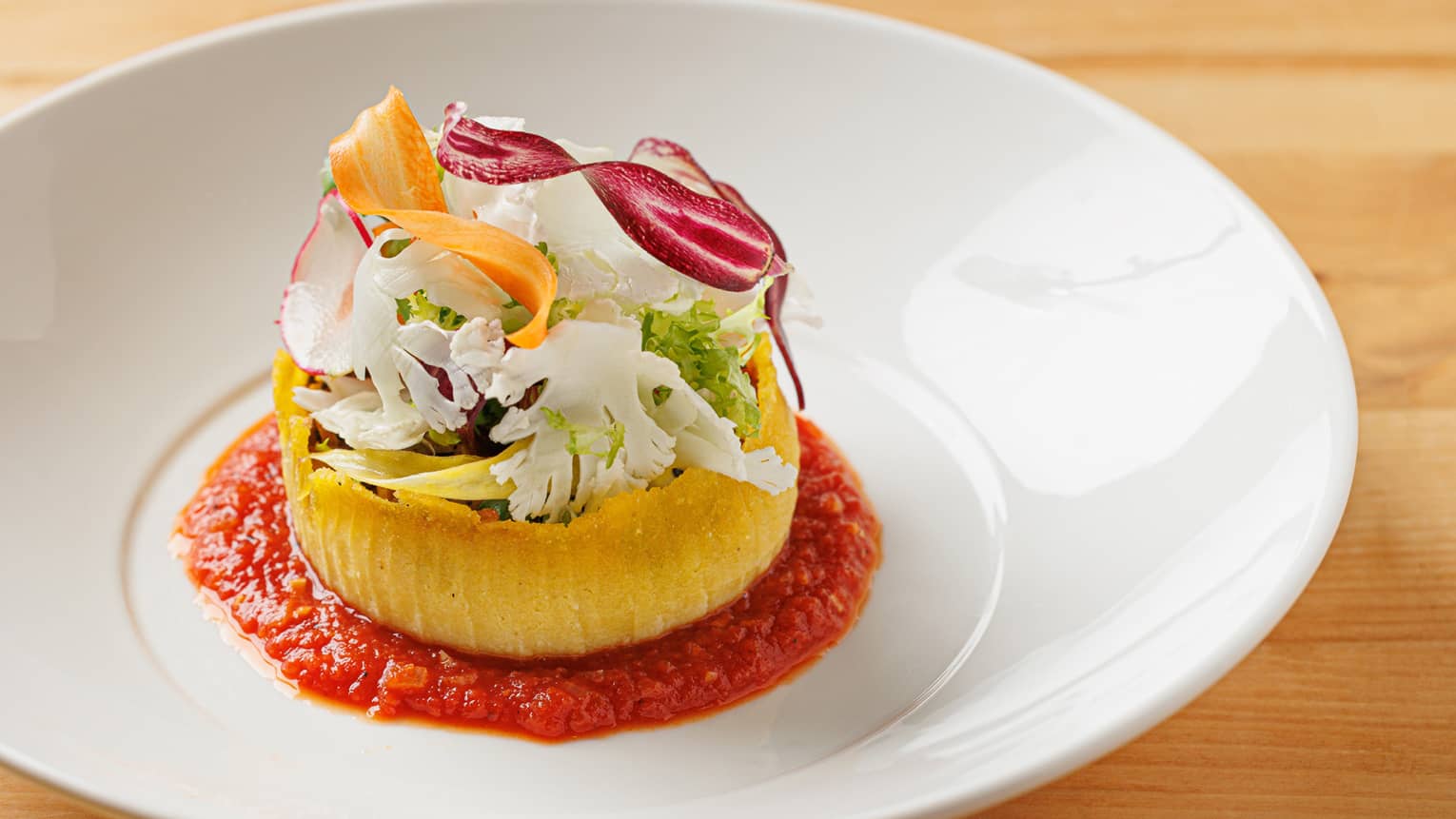Polenta cake topped with shaved vegetables set in marinara sauce in white bowl