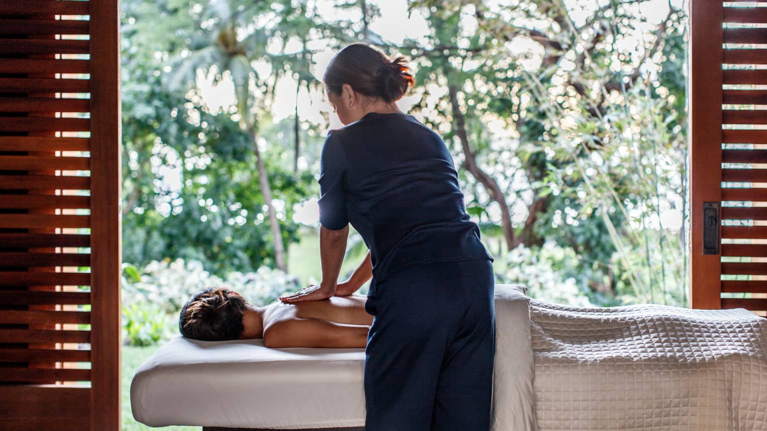 Spa staff massages woman's bare shoulders as she lies under sheet on massage table