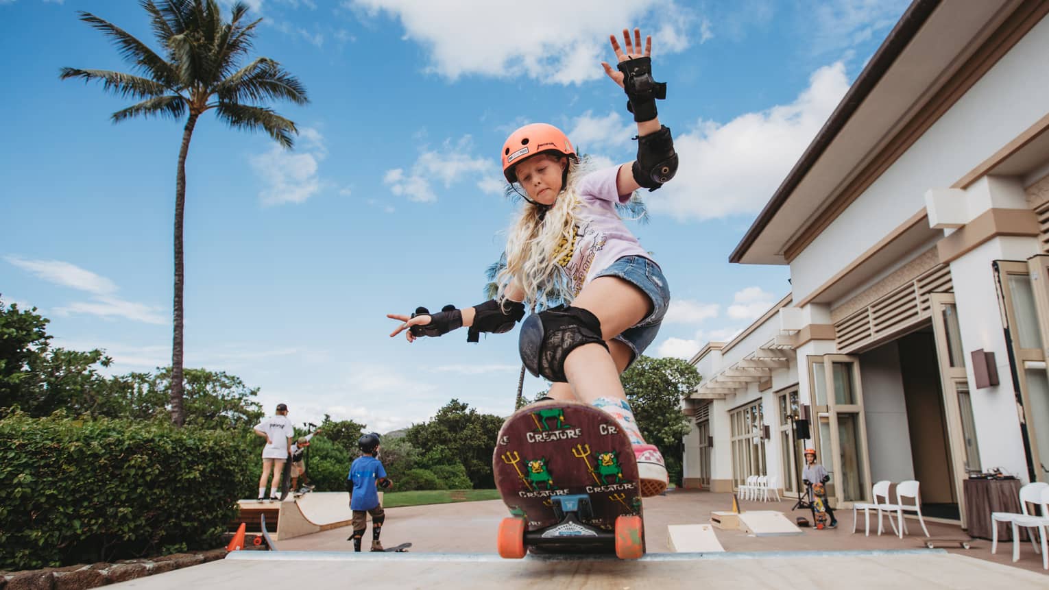 A young girl rides a skateboard at a skate park outside with palm trees